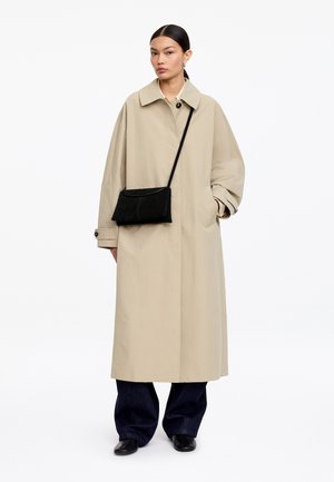 Mujer con un largo abrigo beige tipo trench sobre pantalones oscuros y zapatos negros, con un bolso negro cruzado, de pie frente a un fondo blanco.