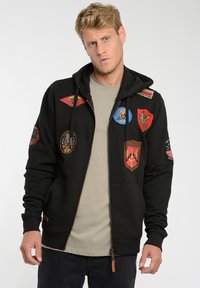 Zwarte hoodie met een rits, voorzien van meerdere kleurrijke patches en badges, een kangoeroezak en een capuchon met trekkoord. Van katoen.