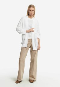 Vit stickad cardigan med intrikata texturerade mönster, i kombination med en lager på lager vit topp och beige löst sittande byxor. Enkla bruna sandaler.