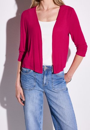 Femme portant un haut blanc avec un cardigan rose vif ouvert sur le devant et un jean taille haute bleu clair, la main reposant dans la poche.