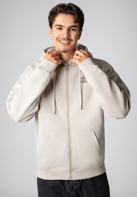 Light beige zip-up hoodie with drawstrings, front pockets, and ribbed cuffs. Smooth texture, relaxed fit, featuring a small logo on the chest.