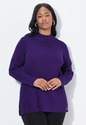 Frau mit mittlerem Hautton trägt einen lila Pullover mit langen Ärmeln und schwarze Hose, steht mit sanft gefalteten Händen vor einem weißen Hintergrund.