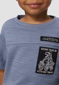 Lichtblauwe korte mouwen shirt met een textuurstof en zwarte patch met witte dinosaurussen en voertuig grafiek, geaccentueerd met tekst.