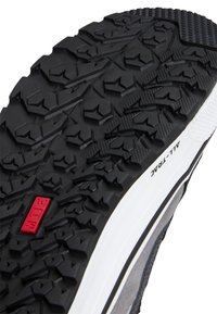 Semelle de chaussure en caoutchouc noir avec un motif de bande de roulement en zigzag profond et en flèche, un logo rectangulaire rouge, et une semelle intermédiaire blanche étiquetée "ALL-TRAC".