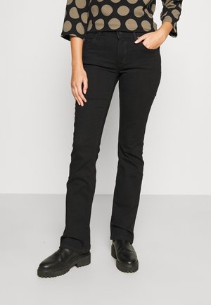 Person wearing black slim-fit jeans, black chunky boots, and a brown top with large gold polka dots, one hand in pocket.