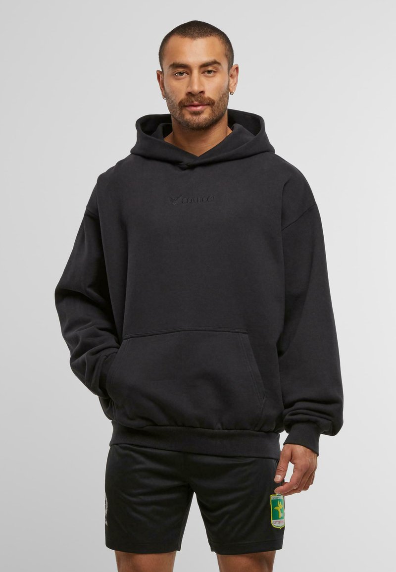 Mann mit kurzem Haar und Bart, der einen schwarzen oversized Hoodie und schwarze Sport Shorts mit einem grünen und gelben Patch trägt.
