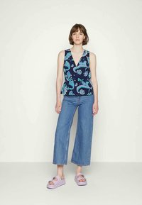 Mulher de pé em calças de ganga largas azuis, top sem mangas azul-marinho com padrão paisley azul, e sandálias de plataforma lilás claras contra um fundo simples.