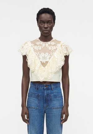 Γυναίκα φοράει κρεμ δαντελένιο crop top με λουλουδάτα σχέδια και βολάν στο μανίκι, συνδυασμένο με ψηλόμεσα μπλε τζιν με μπροστινές τσέπες.