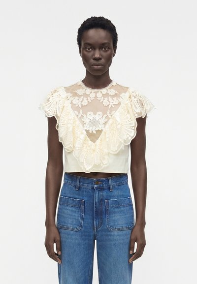 Femme portant un crop top en dentelle crème avec des motifs floraux et des manches à volants, associé à un jean bleu taille haute avec poches plaquées devant.