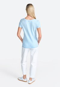Lysblå kortærmet t-shirt med rund halsudskæring, der har en knapdetalje på ryggen. Samlet med hvide bukser og lyse sneakers.
