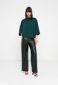 Maglietta oversize verde scuro con collo alto, abbinata a pantaloni larghi in finta pelle nera e sandali con tacco a righe bianche e nere.
