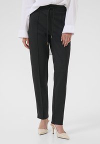 Ženska, oblečena v črne slim-fit hlače s sprednjim šivom in bež koničastimi visokimi petami, v kombinaciji z belo srajco z dolgimi rokavi.
