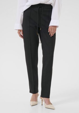 Femme portant un pantalon noir ajusté avec un détail de couture sur le devant et des escarpins beiges à talons pointus, associé à une chemise blanche à manches longues.