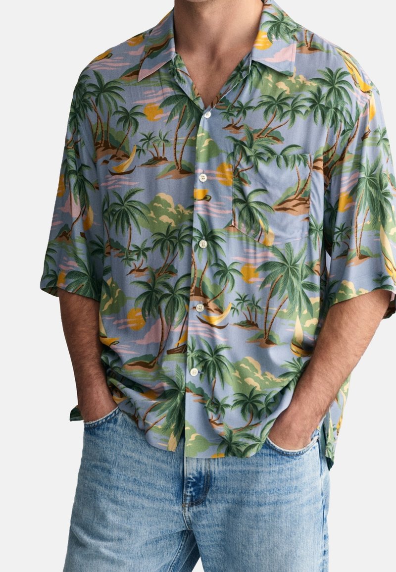 Muž v krátkém rukávu s knoflíky, s tropickým potiskem palm, plachetnic a západu slunce, v kombinaci se světle modrými džínami a rukama v kapsách.