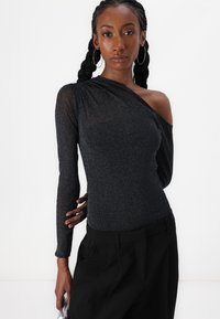 Schwarzer Langarm-Bodysuit aus schimmerndem Stoff mit asymmetrischem Ausschnitt und einer entblößten Schulter, kombiniert mit schwarzen hochgeschnittenen Hosen.