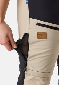 Beiga och svarta utomhusbyxor med dragkedjefickor på sidan och mesh-foder; har en läderetikett med logotyp, tillverkade av slitstift, flexibelt material.