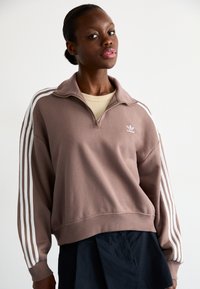 Sweatshirt castanho com fecho éclair e riscas brancas longas nas mangas. Apresenta um corte descontraído e tecido suave. Pequeno logótipo branco no peito.