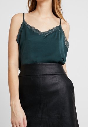 Femme portant un caraco vert foncé orné de dentelle, rentré dans une jupe en cuir noire taille haute, sur un fond uni.