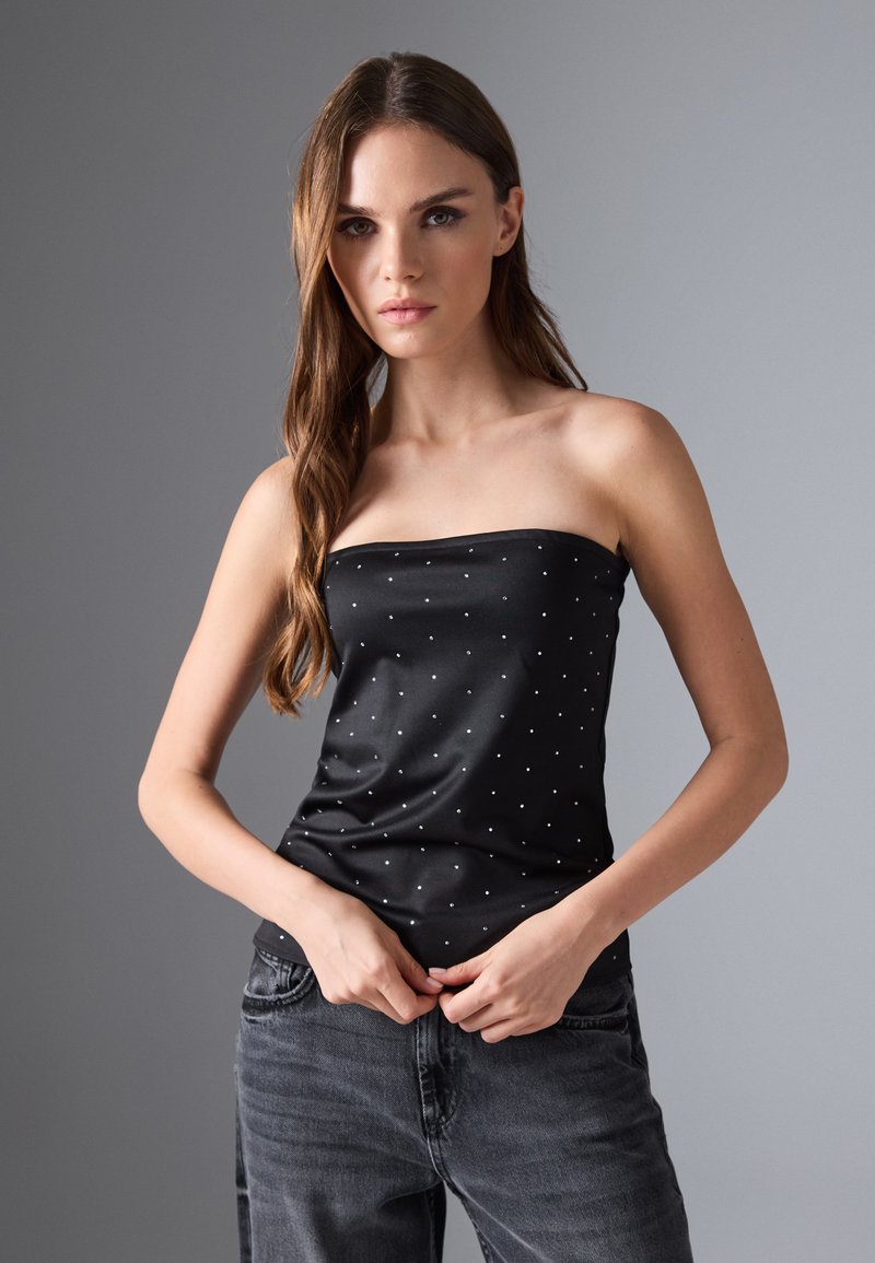 Top negro sin mangas con pequeños lunares blancos, hecho de una tela suave. La modelo lo lleva con unos jeans gris oscuro. Diseño simple y ajustado.