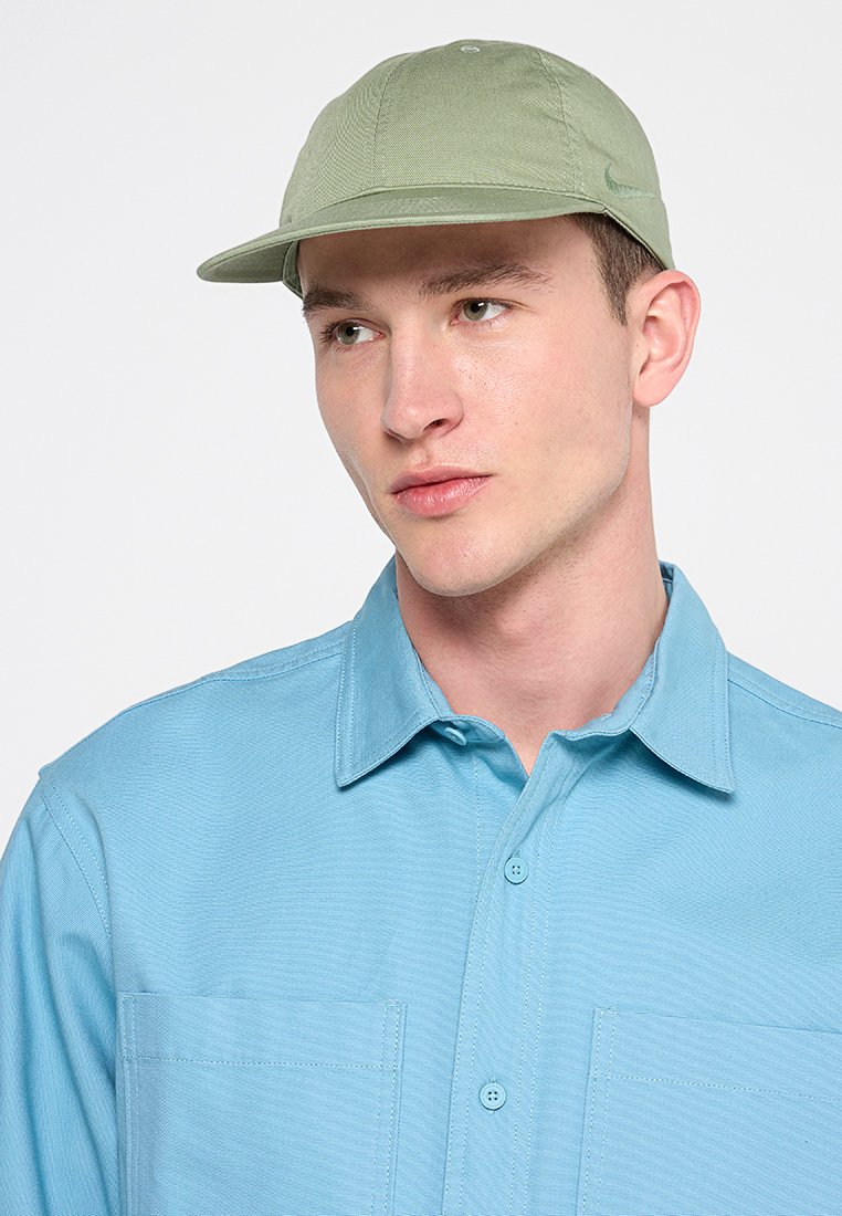 Gorra verde con ala redondeada, hecha de una tela texturizada, llevada sobre una camisa de botones azul claro con dos bolsillos en el pecho.