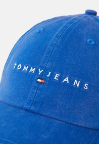 Blue denim baseball cap with white "TOMMY JEANS" text and a rectangular red, white, and blue emblem. Features stitching and a button on top.