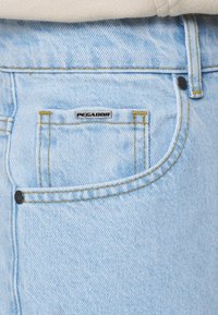 Hellblaue Jeans aus Denim verfügen über eine Vorderentasche mit einem schwarzen "PEGADOR"-Label und sichtbaren gelben Nähten, ergänzt durch einen kontrastierenden schwarzen Knopf.