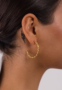 Goldene, verdrehte Creole mit polierter Oberfläche. Kreisform, mittlere Größe, mit einem strukturierten Design, am Ohrläppchen getragen.