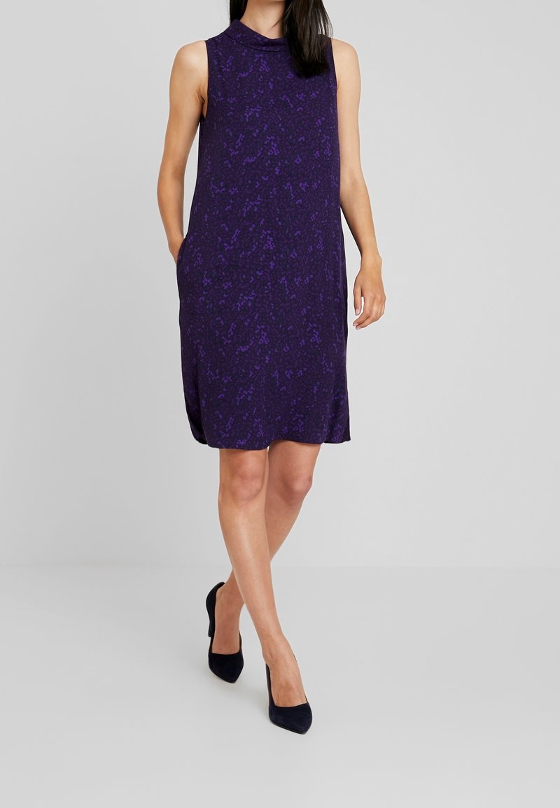 Bez rękawów fioletowa sukienka z fakturowanym wzorem, z bocznymi kieszeniami i wysokim dekoltem. Nosi z czarnymi wysokimi szpilkami.