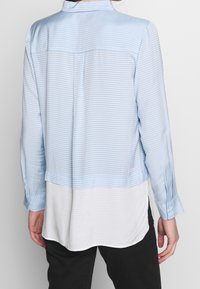 Camicia a maniche lunghe azzurro chiaro con sottili righe bianche, dotata di orlo asimmetrico e chiusura a bottone sulle maniche. Texture del tessuto liscia.