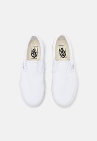 Zapatos slip-on de lona blanca con tejido texturizado, acentos elásticos en los lados y suelas de goma. La marca en las plantillas presenta un logo negro.