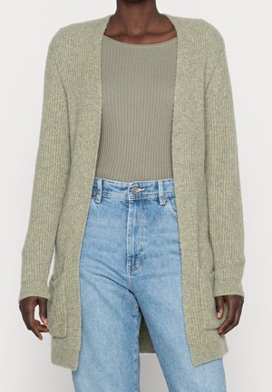 Femme portant un cardigan tricoté vert clair ouvert sur le devant, avec un top côtelé vert olive et un jean bleu clair taille haute.