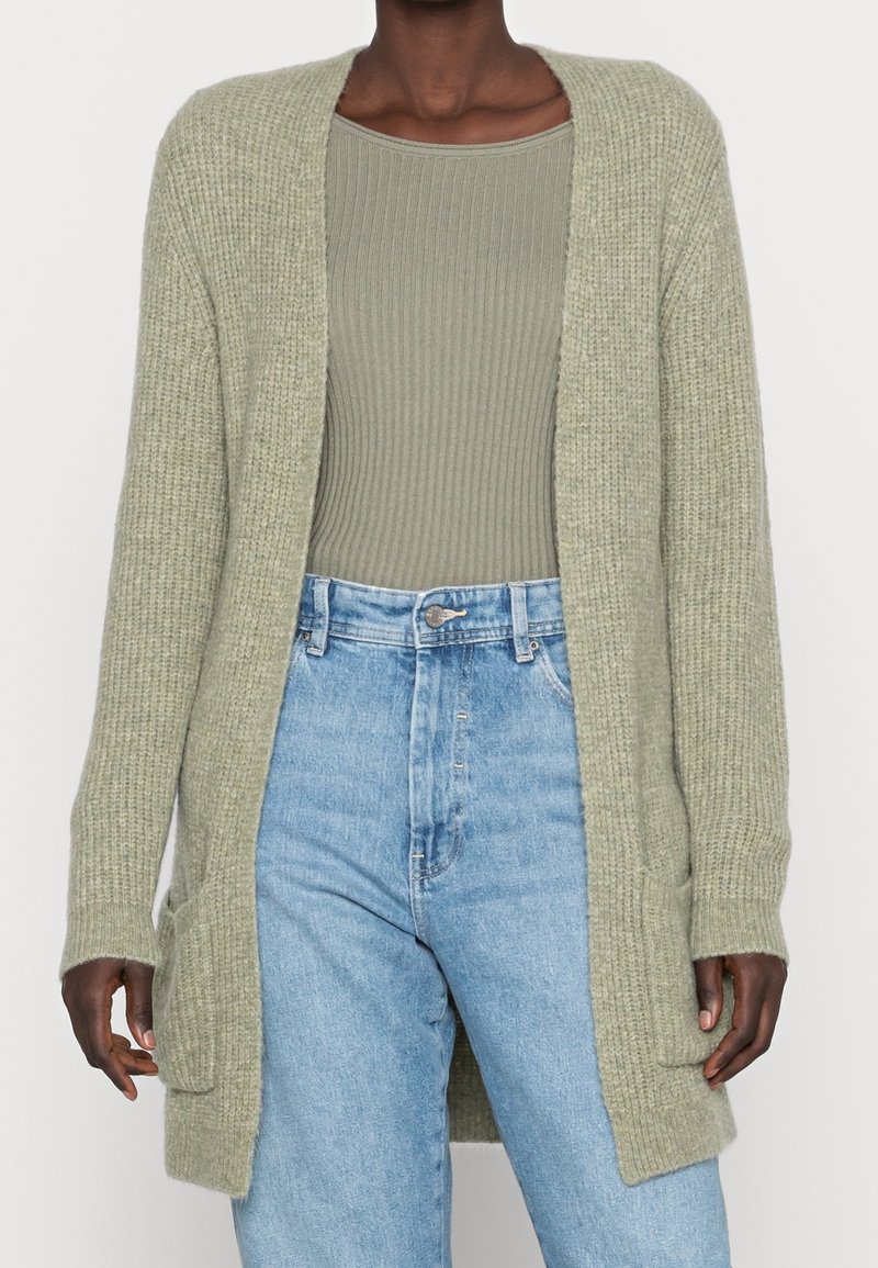 Femme portant un cardigan tricoté vert clair ouvert sur le devant, avec un top côtelé vert olive et un jean bleu clair taille haute.