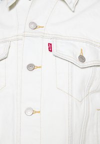 Biała kurtka dżinsowa z srebrnymi guzikami, żółtym przeszyciem, kieszeniami na piersi oraz małą czerwoną metką Levi's na szwie lewej kieszeni.