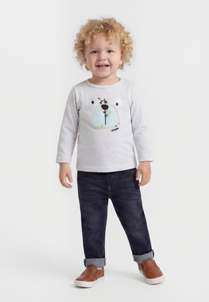 Tout-petit aux cheveux blonds bouclés portant un t-shirt blanc à manches longues avec un motif visage d’ours, jean foncé et chaussures marron sans lacets.