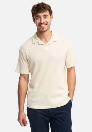 Lachende jonge man met krullend haar, gekleed in een crèmekleurig overhemd met korte mouwen en een kraag, en een donkere broek, staand met één hand in zijn zak.