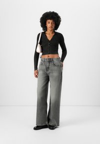 Cárdigan corto de manga larga negro acanalado, jeans de pierna ancha gris claro de cintura alta y calzado negro. La modelo sostiene un pequeño bolso rosa.