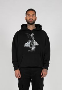 Schwarzer Hoodie mit einem grauen Grafikmotiv eines geflügelten Wesens auf einer Wolke, mit weißem Text unter dem Bild. Aus weichem Stoff mit relaxed Fit.