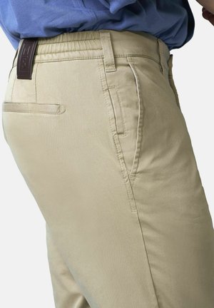 Person, der beige schlanke Hosen mit Seitentaschen und Gesäßtaschen, einem braunen Gürtel mit Schlaufenetikett und einem in die Hose gesteckten blauen Hemd trägt.