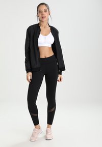 Chaqueta bomber negra, sujetador deportivo blanco y leggings negros con acentos de malla. Zapatillas deportivas de color rosa claro completan el conjunto atlético.