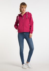Fuchsia lättviktsjacka med huva, justerbara dragsnören, dragkedjestängning och sidofickor, i kombination med blå jeans och vita sneakers.