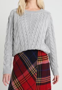 Pull gris clair tricoté avec un motif torsadé, doté d'un col rond et d'une coupe ample. Associé à une jupe à carreaux multicolores.