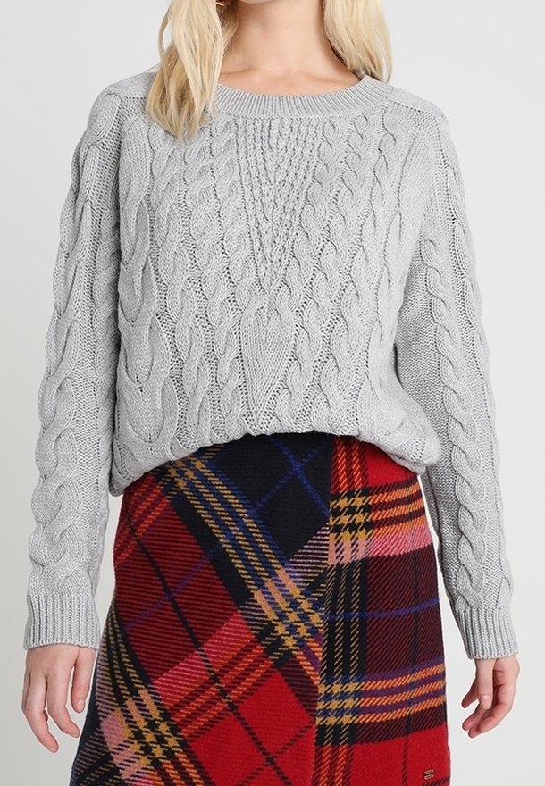 Pull gris clair tricoté avec un motif torsadé, doté d'un col rond et d'une coupe ample. Associé à une jupe à carreaux multicolores.