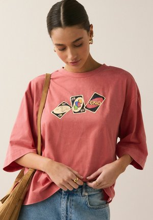 Kvinde iført en oversize lyserød t-shirt med tre trykte UNO-kort, lyseblå jeans, guldsmykker og en brun skuldertaske.