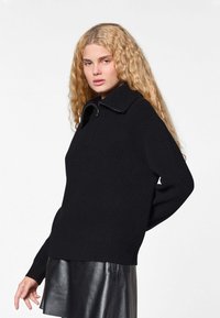 Pull noir en maille côtelée avec un col montant et un détail zippé, porté sur une jupe en cuir noir brillant. Texture lisse, coupe décontractée.