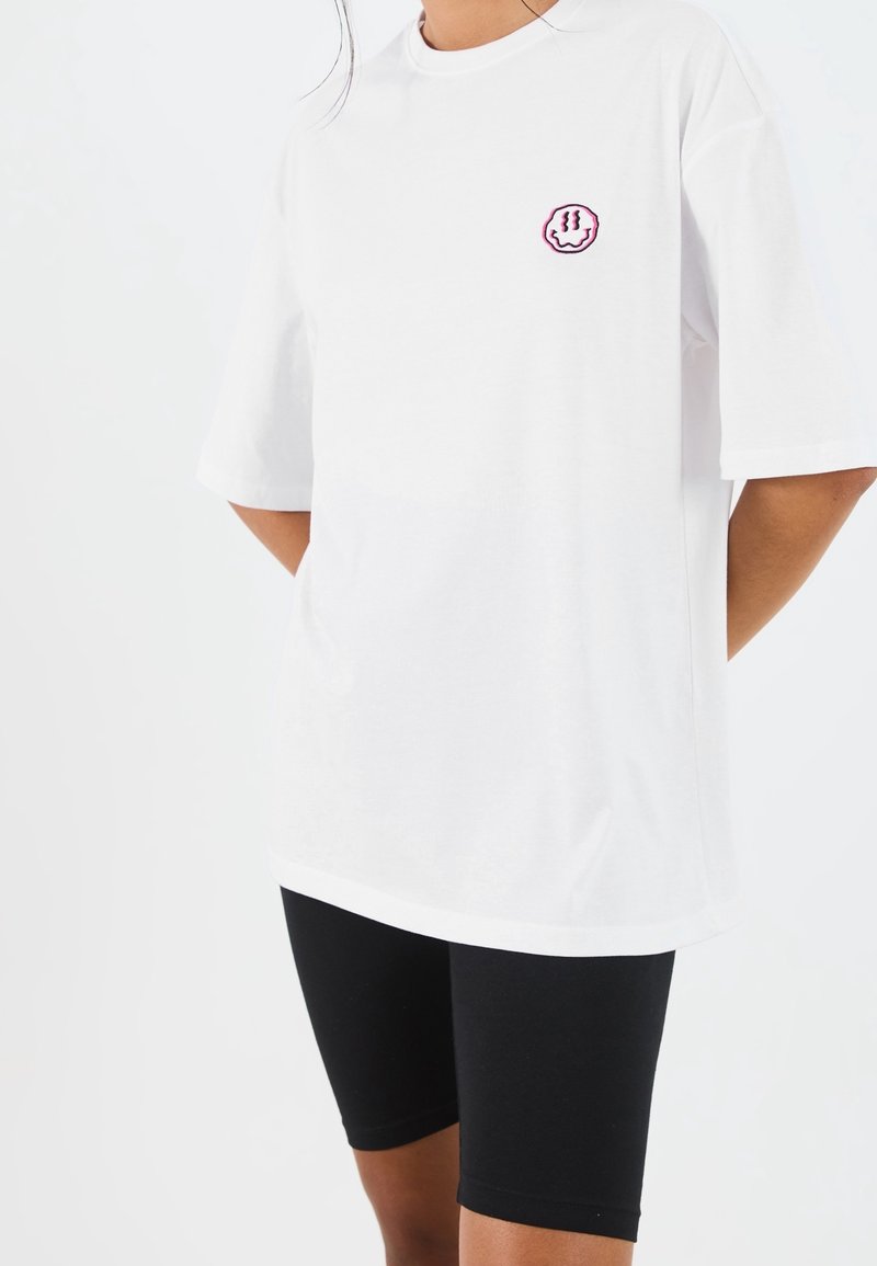 Personne portant un t-shirt blanc oversized avec un petit motif circulaire rose et noir sur la poitrine, associé à un short de cyclisme noir.
