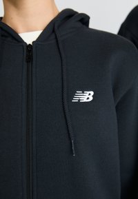 Haut à capuche noir zippé avec une texture douce, présentant un logo New Balance blanc sur la poitrine gauche et des détails de fermeture éclair noirs.