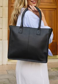 Czarna skórzana torba tote z wytrzymałymi uchwytami, płaskim prostokątnym kształtem, minimalistycznymi detalami szycia oraz małym akcentem logo w dolnym rogu.