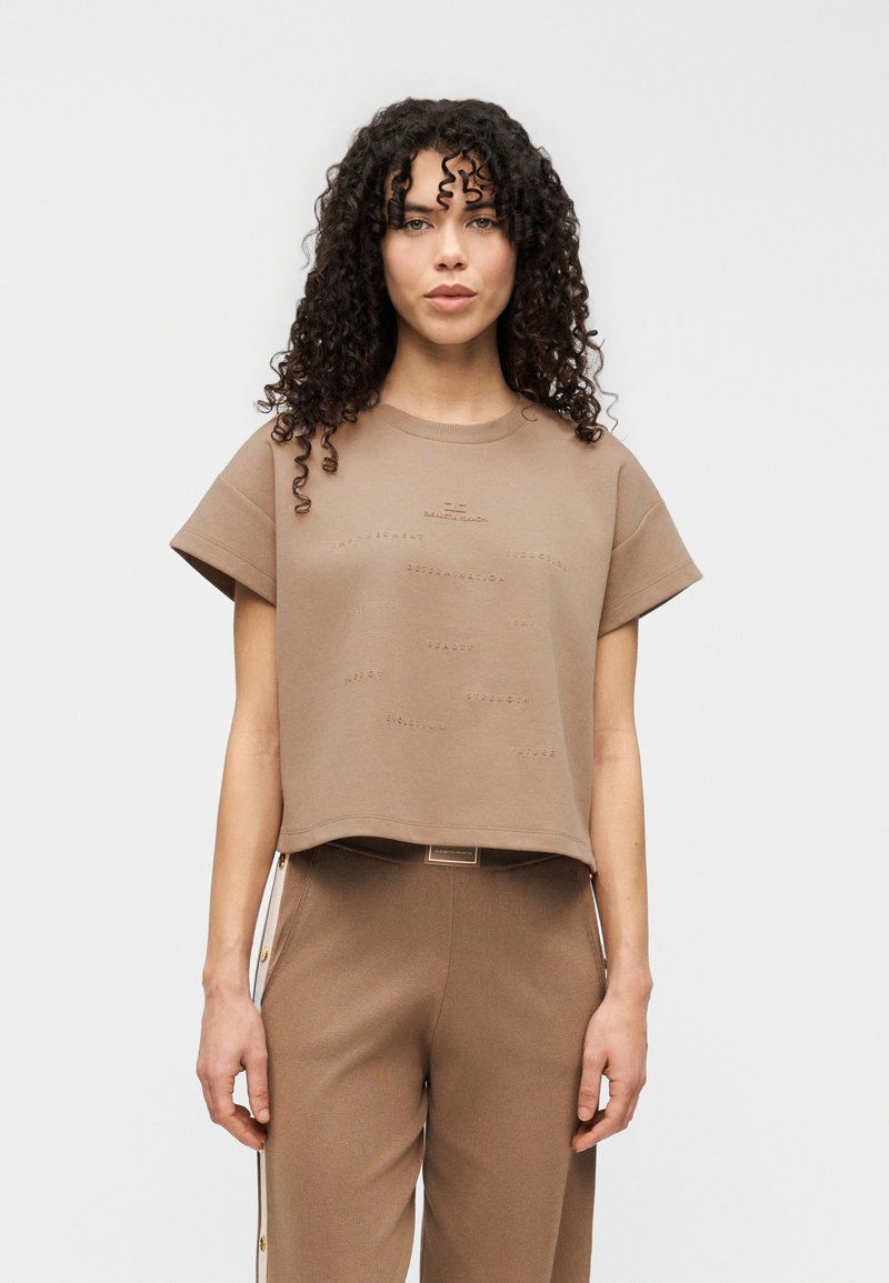 Καφέ κοντομάνικη cropped μπλούζα με ανάγλυφα κείμενα στο ύφασμα. Το ύφασμα είναι λείο, με άνετη εφαρμογή και στρογγυλή λαιμόκοψη.