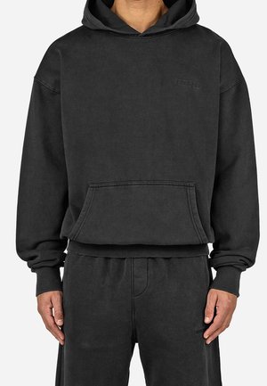 Mężczyzna ubrany w czarną, oversize'ową bluzę z kapturem i kieszenią z przodu oraz dopasowane czarne spodnie, stojący na tle jednolitego białego tła.