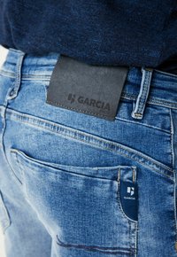 Jeans de mezclilla azul con un parche de cuero negro etiquetado "GARCI A" en la cinturilla. Las características incluyen múltiples bolsillos traseros y costuras visibles.